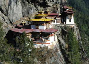 A traditional Bhutanese monastery with white-washed walls and tiered golden roofs, precariously perched on a sheer, vertical grey cliff face overlooking a deep, lush green valley.