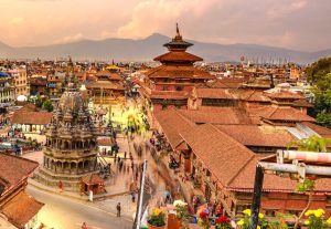 An elevated, wide-angle view of Patan Durbar Square in Lalitpur, Nepal, during sunset, showing the historic Krishna Mandir stone temple, red-tiled pagoda roofs, and the ancient royal palace complex against a backdrop of distant mountains.
