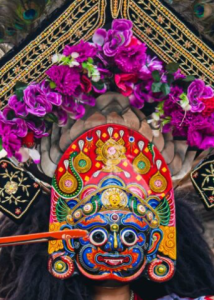 A close-up of a Bhairav mask dancer in Bhaktapur, wearing a vibrant, hand-painted mask and a crown adorned with purple flowers during Gai Jatra.