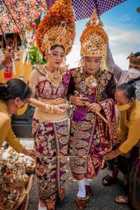 A Balinese couple in elaborate traditional wedding attire known as Payas Agung, featuring gold-plated crowns (gelungan), intricate jewelry, and woven purple and gold sarongs (kamen), during a religious ceremony.