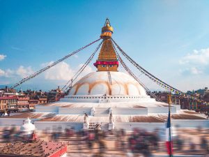 The massive white dome and golden spire of Boudhanath Stupa in Kathmandu, Nepal, adorned with colorful prayer flags under a blue sky.
