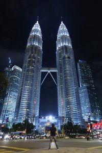 A low-angle shot from a street level in Kuala Lumpur at night. A person stands in the center of the frame, dwarfed by the massive, silver-lit Petronas Twin Towers that dominate the sky above.