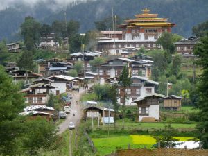 A sprawling view of the Gangteng Monastery, a large white structure with ornate red-and-gold tiered roofs, standing atop a hill overlooking the traditional cluster-style houses of Gangtey village and green fields in a misty mountain valley.