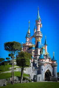 Sleeping Beauty Castle in Disneyland Paris, featuring pink stone walls, blue turrets with gold accents, and manicured square-topiary trees on a grassy hill under a clear blue sky.