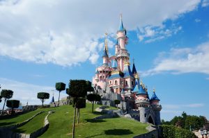 Sleeping Beauty Castle in Disneyland Paris, featuring pink stone walls, blue turrets with gold accents, and manicured square-topiary trees on a grassy hill under a clear blue sky.