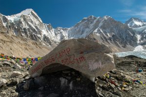 A large, flat rock at Everest Base Camp in Nepal with "EVEREST BASE CAMP 5364m" spray-painted in red, surrounded by colorful prayer flags and snow-capped Himalayan peaks under a clear blue sky.