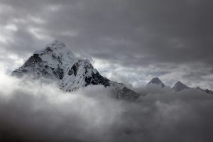 A majestic snow-covered pyramidal mountain peak rising above a dense layer of dark, dramatic clouds in a black and white Himalayan landscape.