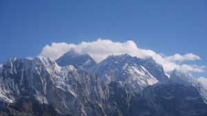 A panoramic view of the snow-covered Himalayan mountain range under a clear blue sky, featuring the dark summit of Mount Everest partially shrouded in white clouds, flanked by the massive Nuptse ridge to the right.