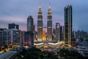 A high-angle city view of Kuala Lumpur during the blue hour. The Petronas Twin Towers are the centerpiece, flanked by other modern skyscrapers and a park with a colorful fountain at the base.