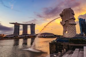 A side profile of the Merlion statue spouting water into Marina Bay during a vibrant orange and purple sunset. In the background, the three towers of the Marina Bay Sands hotel stand prominently against the glowing sky.