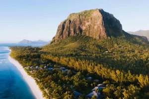 Aerial view of Le Morne Brabant, a large basaltic mountain on the lush green peninsula of Mauritius, bordered by a white sand beach and turquoise ocean water.