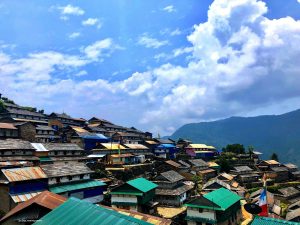 A high-angle landscape photo of a sprawling mountain village in Nepal. Numerous small houses with slanted roofs are built close together on a steep green slope. While many have traditional stone roofs, several feature bright green or blue metal roofing. In the background, a large green mountain is partially obscured by clouds under a bright blue sky with white, puffy clouds. A small Buddhist prayer flag is visible in the lower right foreground.