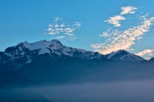 A wide landscape photo of jagged, snow-covered mountain peaks in Nepal under a clear blue sky with light, wispy clouds. The lower third of the image is filled with a soft blue morning mist or fog, partially obscuring the dark foothills.