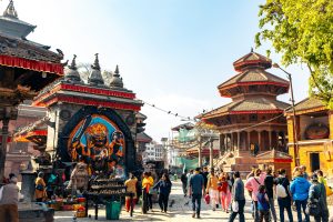 A sunny, wide-angle photo of Bhaktapur Durbar Square in Nepal, featuring ancient brick temples in Shikhara and Pagoda styles, a paved square with several tourists and locals, and traditional Newari architecture.
