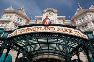 The ornate green iron entrance gate of Disneyland Park in Paris, featuring a large "Disneyland Park" sign in gold and red, with the pink Victorian-style Disneyland Hotel and its clock tower in the background.