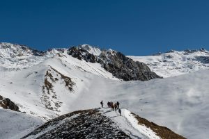 A group of trekkers stands on a snowy mountain ridge in Nepal's Langtang Valley, with a clear blue sky overhead and large, snow-capped peaks in the background. 