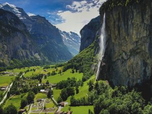 A dramatic high-angle view of a massive waterfall cascading down a sheer vertical cliff into a lush green Alpine valley featuring a small Swiss village and snow-capped peaks in the distance.