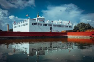 A horizontal view of the white Maya Devi Temple in Lumbini, Nepal, reflected in the Puskarini holy pond, with the Ashoka Pillar visible in the background under a blue sky with white clouds.