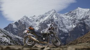 A camouflage-patterned Royal Enfield Himalayan motorcycle parked on a high-altitude dirt trail with massive snow-capped mountains in the background.