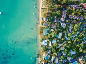 Bird's-eye view of a coastal tropical resort area in Mauritius, featuring white-roofed buildings nestled among lush green palm trees next to a turquoise ocean with several small boats. 