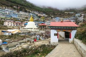 A white Buddhist stupa with a gold spire stands beside a traditional red-roofed entrance gate in the mountain village of Namche Bazaar, Nepal.