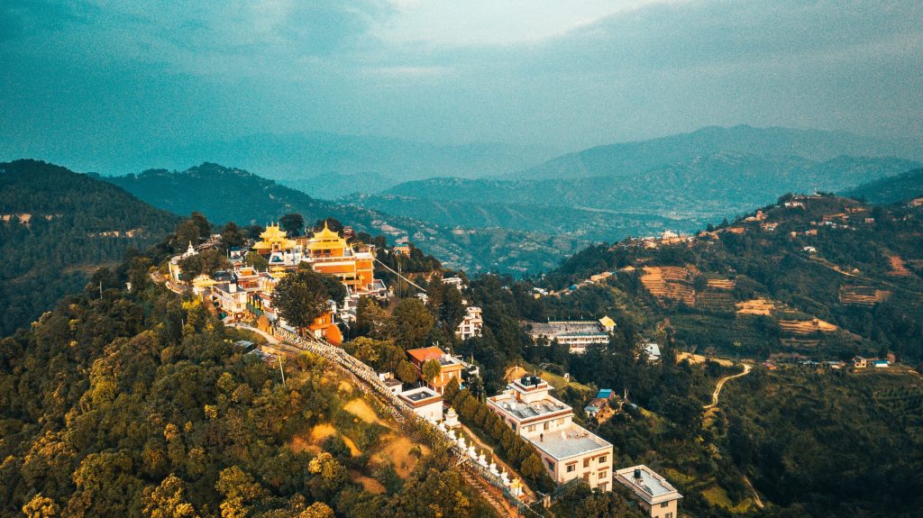 Aerial panoramic view of Namobuddha Monastery (Thrangu Tashi Yangtse) on a forested hilltop in Nepal, featuring golden-roofed temple buildings, a long stairway lined with white stupas, and the rolling green hills of the Kavre region under a soft twilight sky.