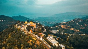 Aerial panoramic view of Namobuddha Monastery (Thrangu Tashi Yangtse) on a forested hilltop in Nepal, featuring golden-roofed temple buildings, a long stairway lined with white stupas, and the rolling green hills of the Kavre region under a soft twilight sky.