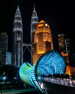 A night shot featuring the brightly lit, colorful Saloma Link pedestrian bridge in the foreground. The bridge’s intricate blue and green LED patterns lead the eye toward the Petronas Twin Towers in the background.