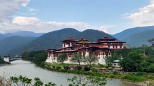 A massive white fortress with tiered red and gold roofs, known as Punakha Dzong, situated at the edge of a wide river with green hills and misty mountains in the background under a cloudy blue sky. A traditional cantilever bridge is visible to the left.