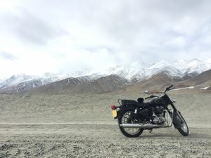 A classic black Royal Enfield Bullet motorcycle parked on a vast, rocky desert plain with snowy mountain ranges in the distance.