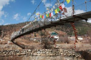 A long, traditional iron chain suspension bridge adorned with colorful Buddhist prayer flags spans a dry riverbed in a hilly region of Bhutan.