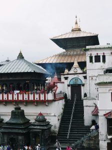 Multi-tiered pagoda-style temple complex in Nepal with worshippers standing along a red-and-white balcony, smoke rising from incense, and stone shrines in the foreground beneath an overcast sky.