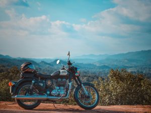 A Royal Enfield Classic motorcycle parked on a hilltop dirt road with a scenic valley and blue sky in the background.