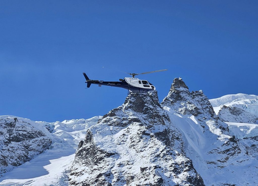 A helicopter hovering or landing on the tip of a steep, rocky, and snow-covered mountain peak against a clear blue sky.