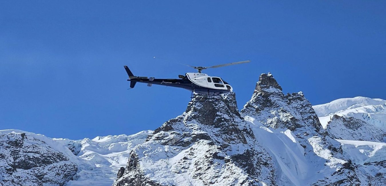 A helicopter hovering or landing on the tip of a steep, rocky, and snow-covered mountain peak against a clear blue sky.