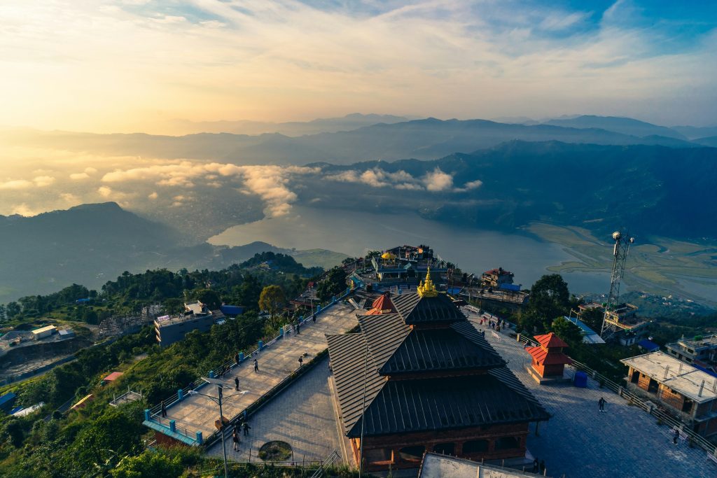 An aerial photo of the Panchamukhi Ganesh temple at Sarangkot hill, Pokhara, Nepal, at sunrise, with a lake and mountains in the misty background.