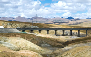 Long railway bridge crossing a desert valley in the Tibetan Plateau with mountain ranges in the background under a cloudy sky.