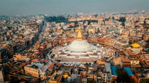 Aerial view of the large, white Boudhanath Stupa with a golden spire, surrounded by a dense urban area in Kathmandu, Nepal.