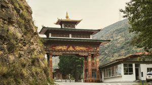 A large, ornate traditional Bhutanese gateway with three-tiered golden roofs and intricate carvings, standing across a highway road at the entrance to Thimphu, with steep rocky hills in the background.