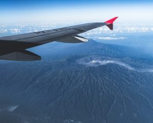 A view from an airplane window showing a grey wing with a red tip flying over a massive, dark volcanic crater in a mountainous landscape.