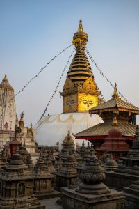A vertical shot of the golden Swayambhunath Stupa (Monkey Temple) in Kathmandu, Nepal, surrounded by smaller stone chaityas and pagodas, with the all-seeing Buddha eyes and colorful prayer flags against a clear sky.