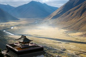 A traditional Tibetan building with a tiered roof overlooks the mist-covered Yarlung Valley with a winding river and surrounding mountains in Tibet.