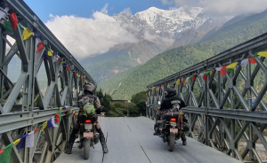 Two adventure motorcyclists on dual-sport bikes crossing a metal Bailey bridge adorned with colorful Tibetan prayer flags, heading toward a massive snow-capped Himalayan peak.