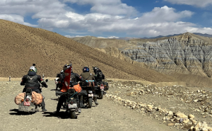 A convoy of adventure motorcyclists riding on a dirt trail through the arid, rolling hills and stratified rock formations of the Upper Mustang desert under a blue sky.