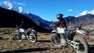 Two white Royal Enfield Himalayan motorcycles parked on a grassy ridge overlooking a deep valley and massive snow-capped mountains under a clear blue sky in Nepal.