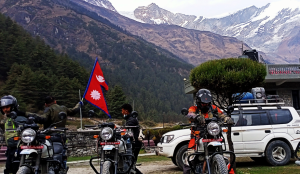A convoy of four adventure motorcyclists riding on a winding dirt track through the arid, high-altitude desert of Upper Mustang, Nepal, with a rider prominently flying the Nepal national flag.