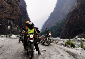 Motorcyclists wearing high-visibility vests riding on a narrow, rocky dirt road through a deep mountain gorge with steep cliffs and a riverbed in the background.