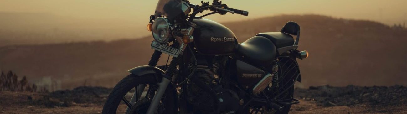 A black Royal Enfield motorcycle parked on a dirt ridge overlooking a hazy mountain valley during sunset in the Himalayas.
