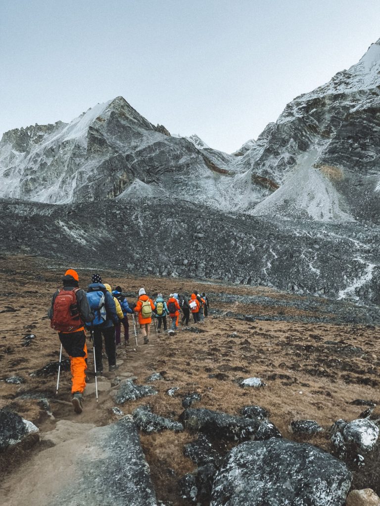 A group of adventure trekkers wearing colorful winter gear hiking along a rocky trail toward snow-capped Himalayan mountains in Nepal.