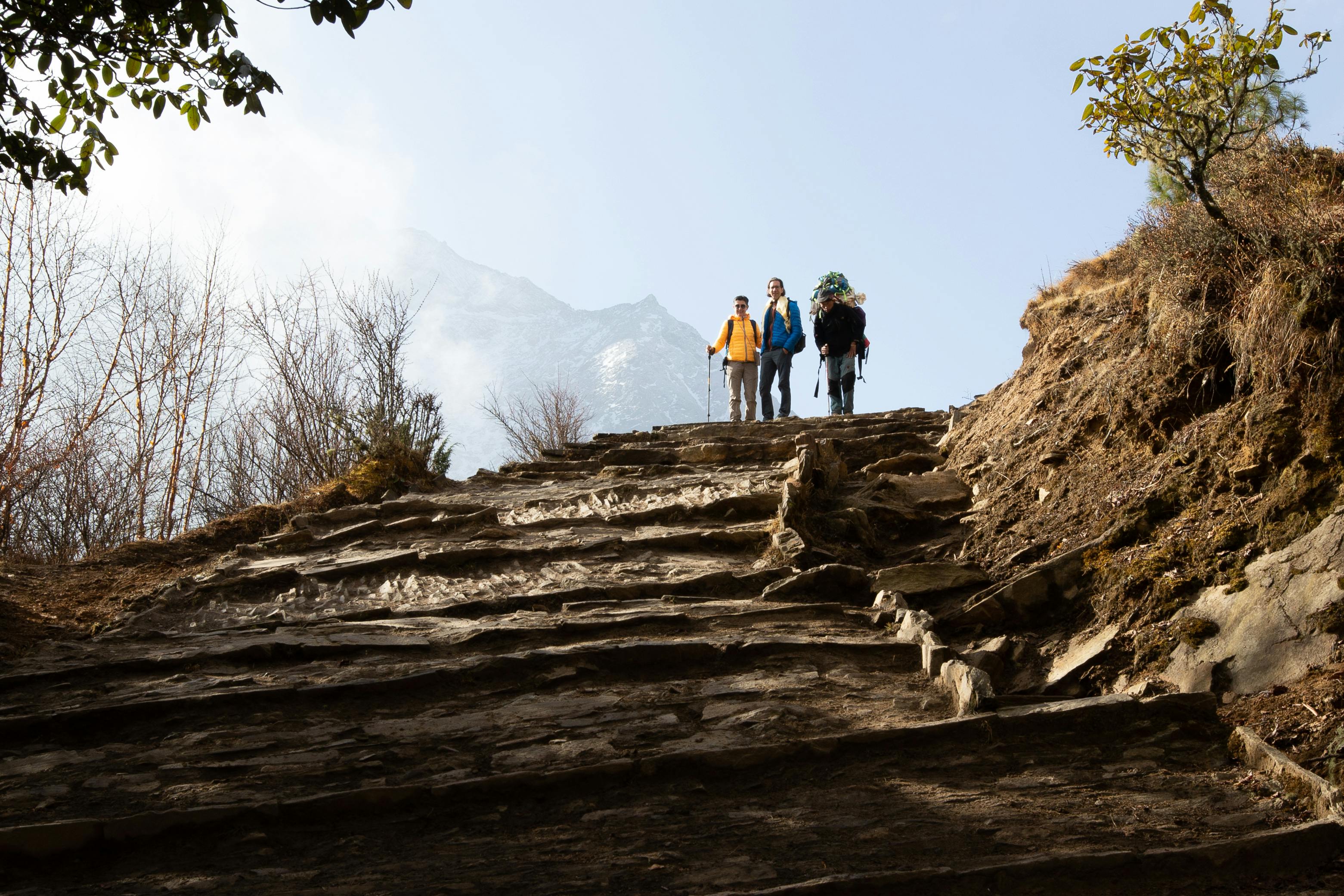 Three trekkers with backpacks and poles ascending a steep stone staircase on a mountain trail in Nepal with a snow-capped peak in the background.